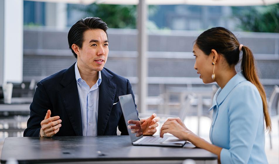 A man and a woman sit outside in a business meeting. The woman has her laptop and the man is dressed in a suit and shirt. 