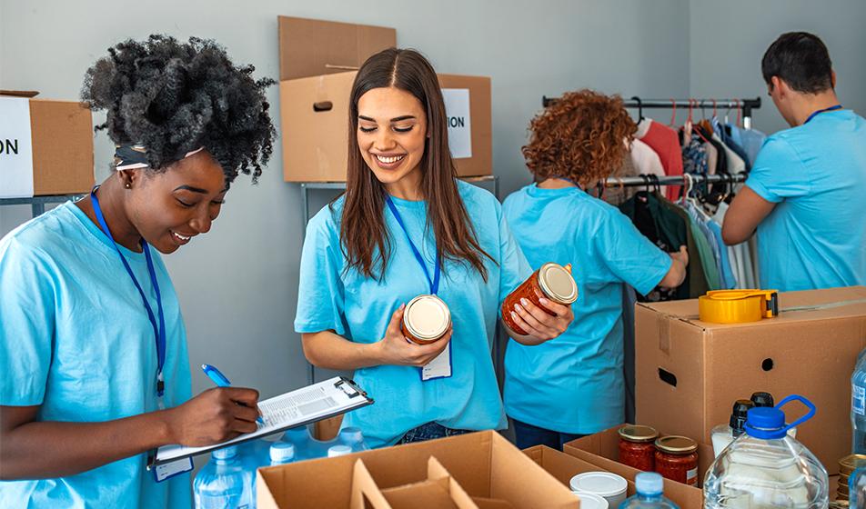 A group of 4 volunteers are packing boxes of food donations and sorting clothes for a charity. They are all wearing blue t shirts