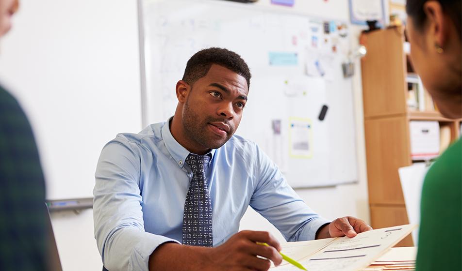 A man sits at a desk across from a man and a woman, who can only be slightly seen at the edge of the image, out of focus. The man has a piece of paper that he's pointing to with a pen. His expression is serious. 