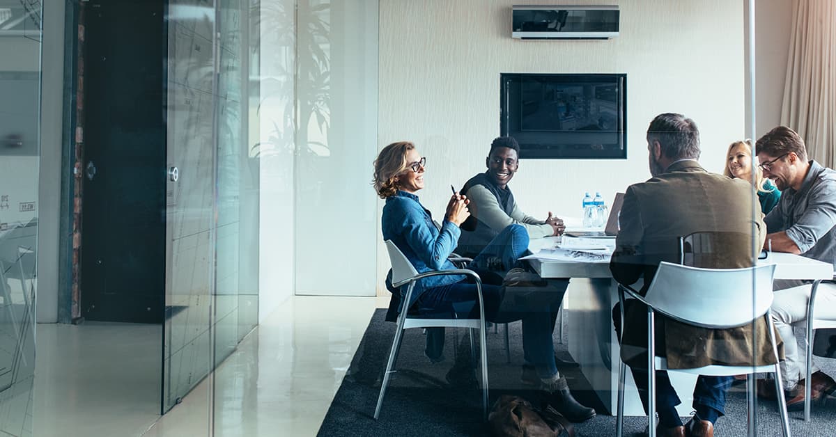 Five people enjoying conversation, sat around a white table within a glass-walled office.