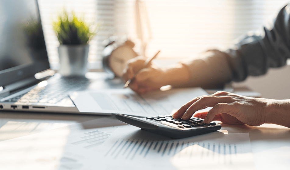 A person sat at a desk with their laptop and a piece of a paper. The person is using a calculator to complete their work.