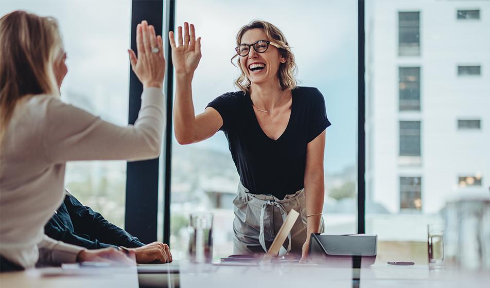 Two women high-five across an office desk. The woman facing the camera is smiling widely, looking happy. 