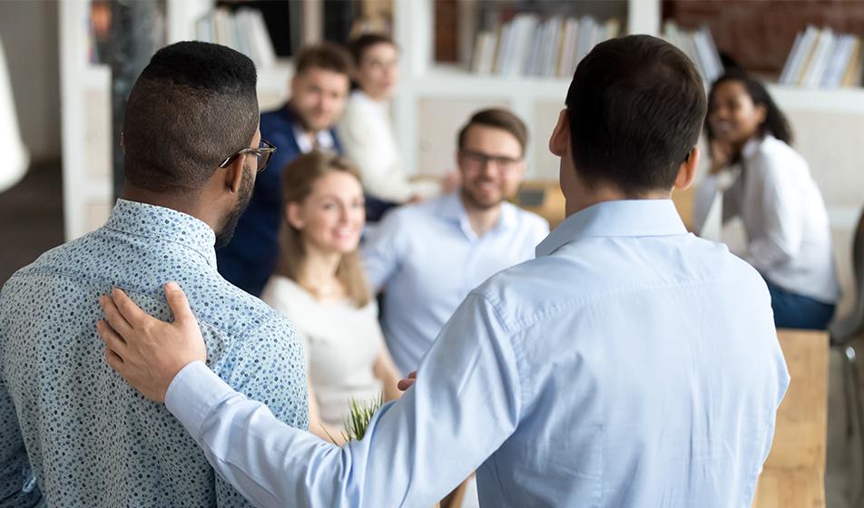 A new team member is being welcomed to their smiling team. Two men stand with their backs to the camera, one has their arm on the other's shoulder, in a supportive gesture.