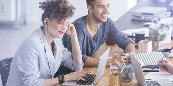 A woman and a man sat working at a desk together. The woman has her headphones in and is working on her laptop. The man is sat next to her and is talking to a colleague across from him.