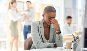 A woman sits at her desk. Her eyes are closed, and she's pinching the bridge of her nose with one hand. She looks stressed. People stand and talk in the background, out of focus. 