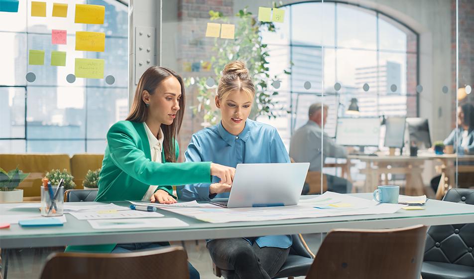 Two women colleagues sit in a modern office with glass walls. One is pointing at the other's laptop, explaining something.