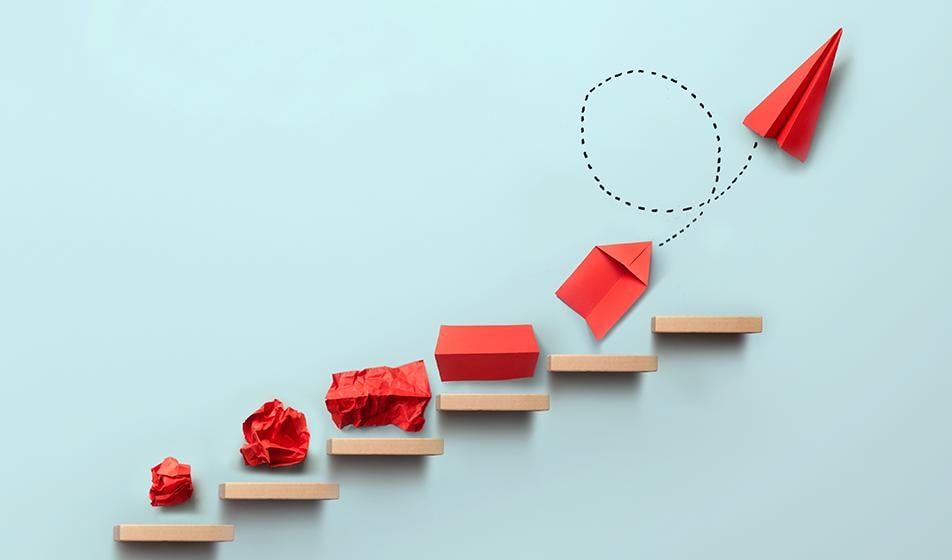 Floating wooden shelves are displayed on a pale blue background, in gradually ascending order up to the right. On each shelf is a piece of red paper - on the first shelf it's crumpled into a ball, and the final highest shelf it's a paper aeroplane. The between shelves show the gradual unfurling into the paper aeroplane.