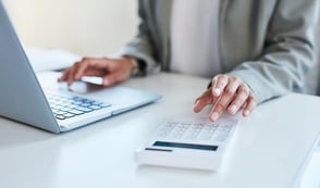 Someone is at a desk with one hand on a laptop and with one hand typing into a calculator. Their hands are the focus of the image. 