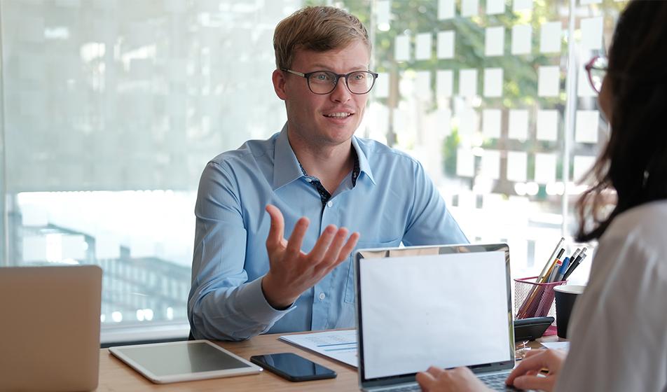 A man with glasses sits across a desk from his manager, a woman, with a laptop open. He's gesturing with his hands as if he's explaining something. 