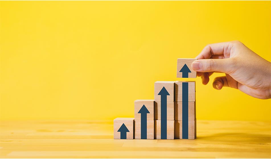 A yellow background shows an abstract image of wooden cubes showing upward-pointing arrows, increasing in height. A hand places the final arrowhead block on the tallest cube pile on the right of the image. 