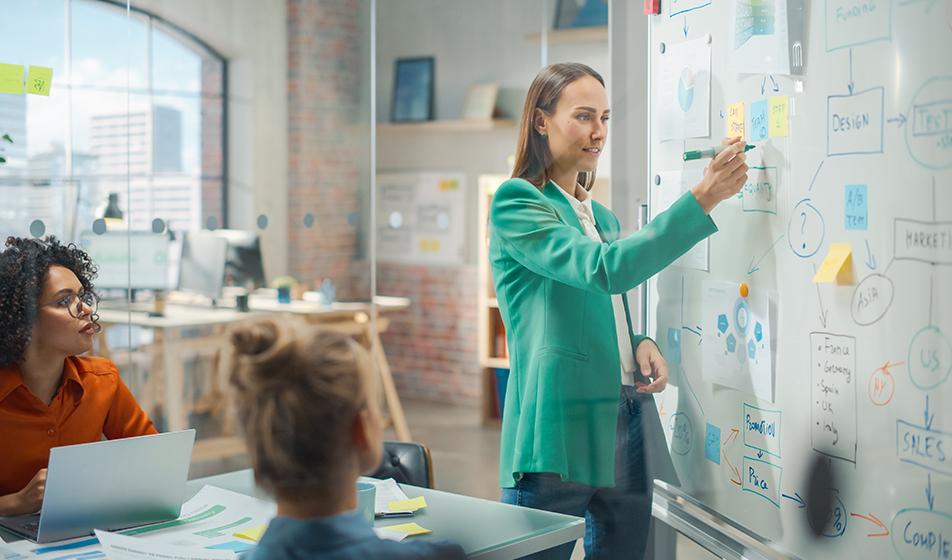 In an office, a woman is plotting a mind-map chart on a whiteboard. Another woman sits at a desk nearby, looking at the board. 