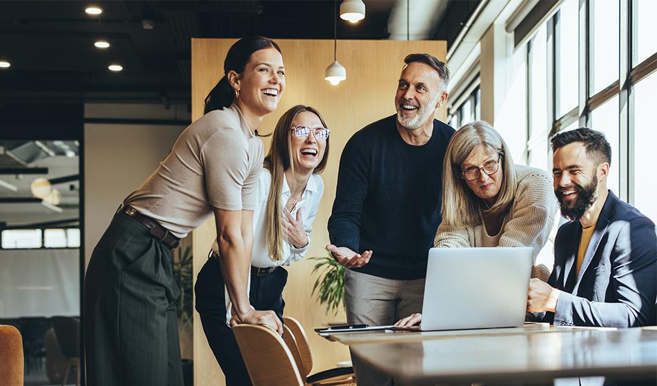 A group of people huddle around a laptop in an office. They're all smiling. 