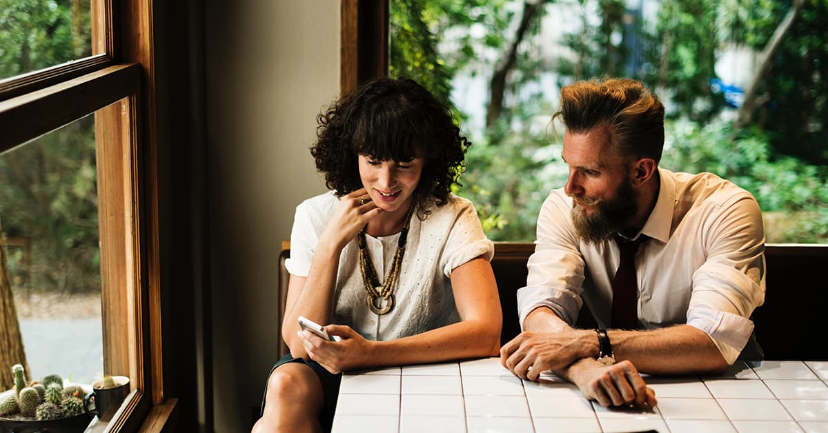 A man and a woman are sat at a wooden table together. They are both wearing white shirts. The woman is sat closest to the window and is showing the man something on her phone. On the window ledge there are some cacti. 