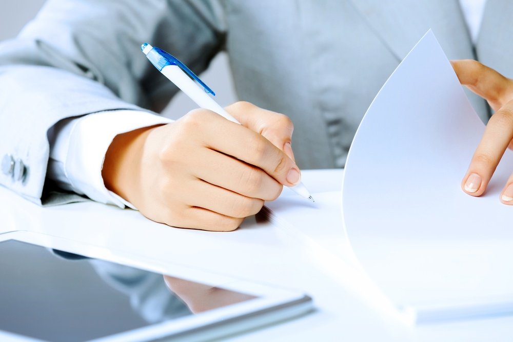 A person in a grey suit is writing on a piece of paper with a blue and white pen. Lying next to the paper is the screen of a tablet.