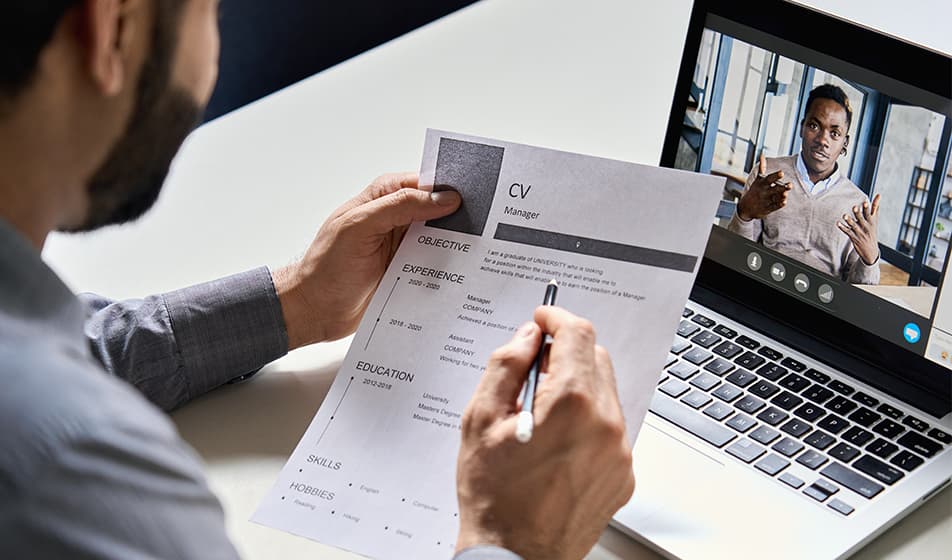 A video interview takes place. The recruiter looks at the interviewee on their laptop screen, whilst holding a printed copy of their CV in their hand. 