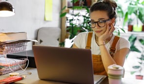 A woman wearing a white shirt and yellow dungarees is working from home. The woman is sat at her desk with a laptop in front of her. Her laptop is surrounded by paperwork, her phone, a cable and a coffee cup. Behind her is a shelf full of plants.