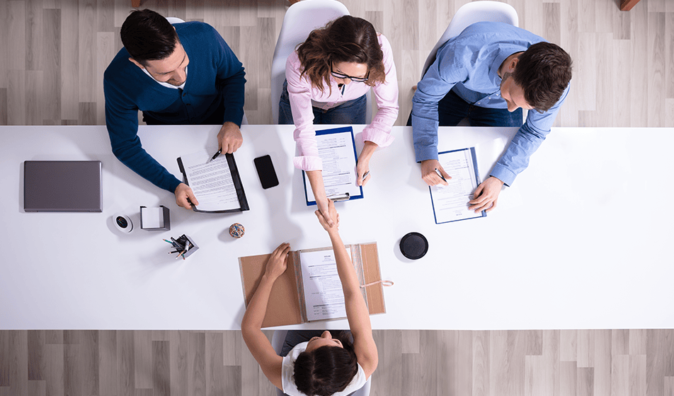 Four people are sat at a white table. Three people are at one side of the table and one person is sat alone on the other side. The person in the middle of the three colleagues is shaking the person's hand across from her. The camera view is looking down from above.