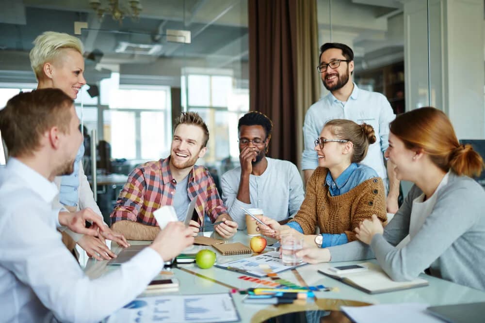 Seven colleagues are gathered around a table in one of their office rooms. They are all laughing with each other. The table is covered in paperwork and stationery.