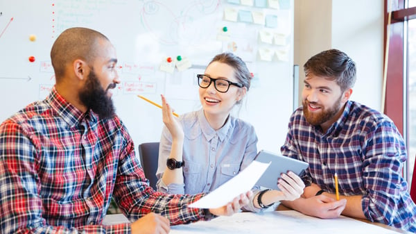 Three colleagues are sat working at a desk together. Two of them are men wearing flannel shirts and one is a woman wearing a blue shirt and glasses. They are laughing in the office.