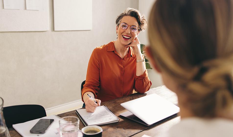 A woman sits at her desk, smiling. The back of another woman's head is shown, facing her. 