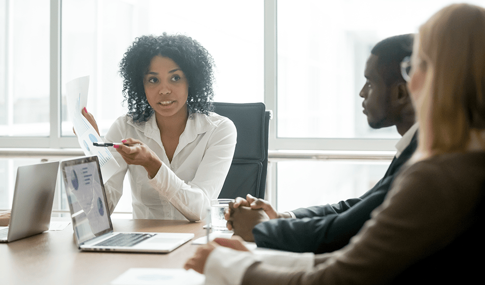 Three colleagues sat down together at a table having a discussion. On the table is a laptop showing various bar charts. 