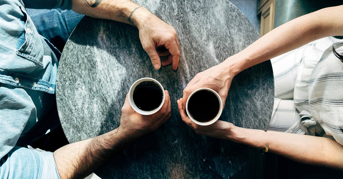 The camera is pointing down on two people sat at a small round table drinking coffee together.