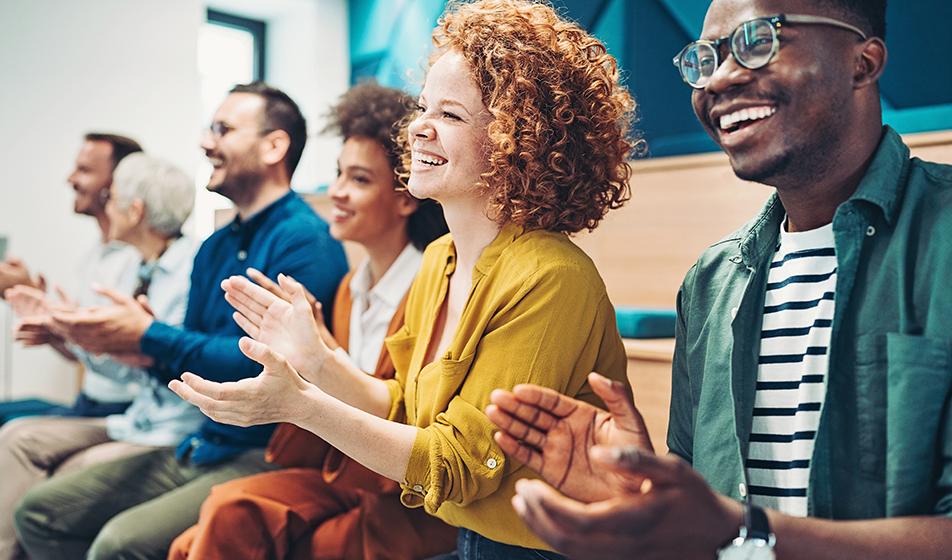 A diverse group of smiling employees are clapping, applauding someone in what looks like a presentation. 