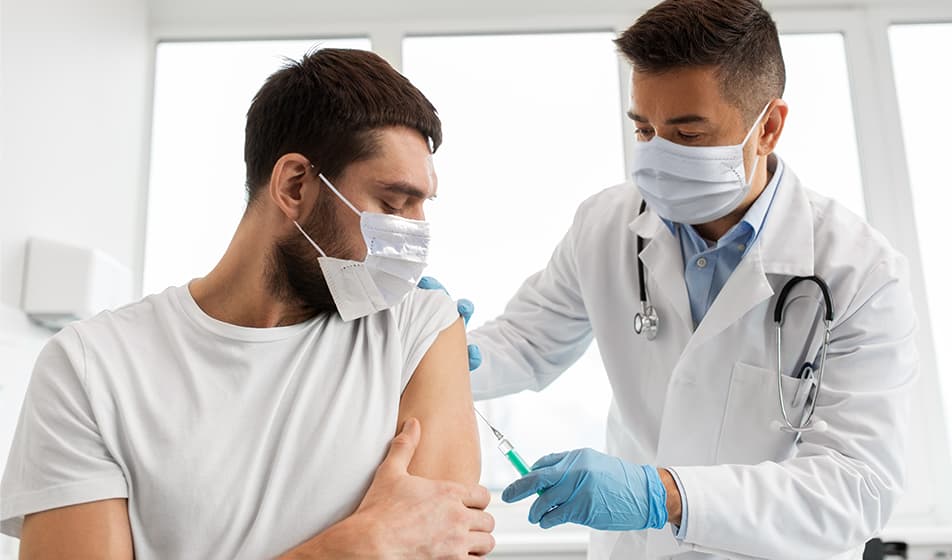 A man in a white t-shirt is wearing a mask and receiving his COVID-19 vaccine from a doctor.