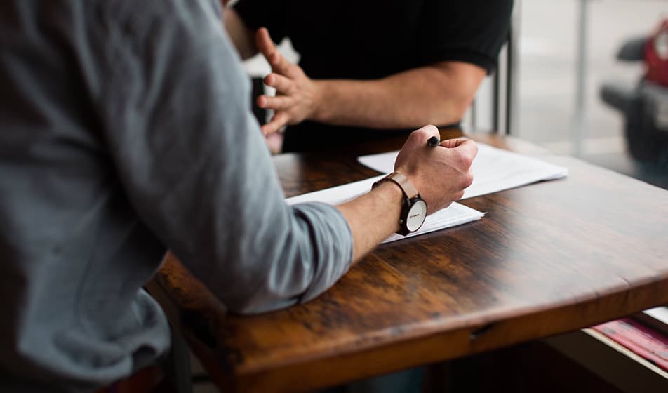 Two men are having a conversation at a wooden table. One man is wearing a grey shirt and is writing notes as he listens to the other man.