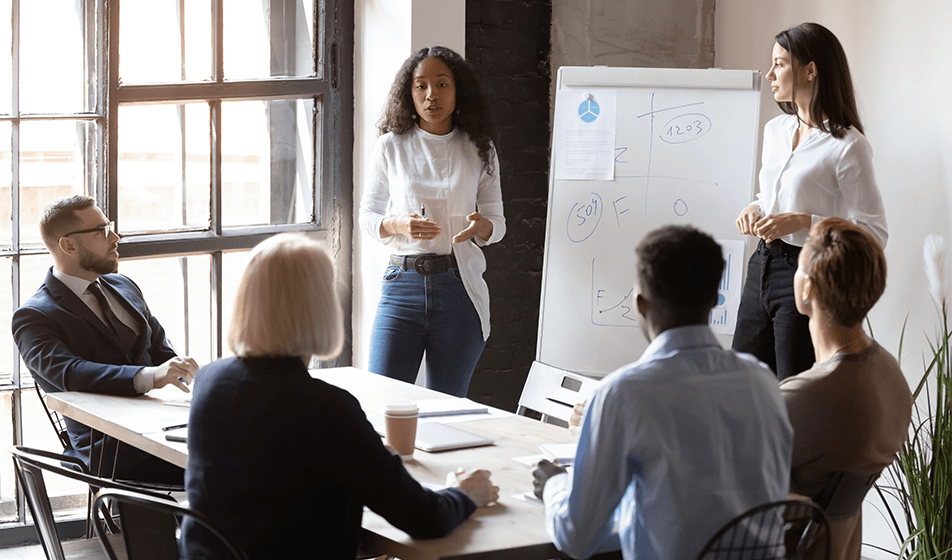 Two women stand in front of a few employees who are seated. They are looking at a board and the women appear to be offering coaching or training. 