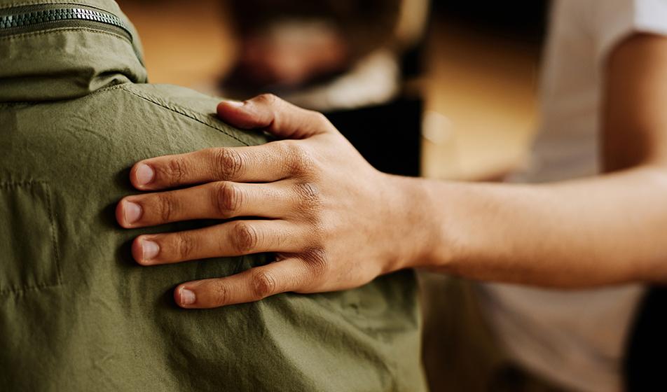 Someone places their hand on another's shoulder in a supportive manner. In the background, other people appear to sitting on chairs in a circle, out of focus.