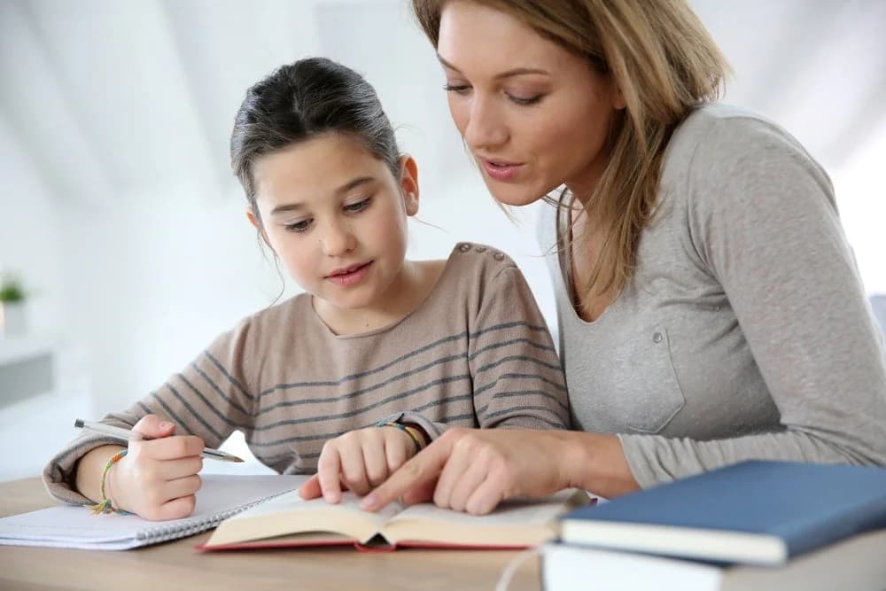 A woman and her daughter are sat at a desk together. The mum is pointing at a page in a notebook and helping her daughter with her homework.