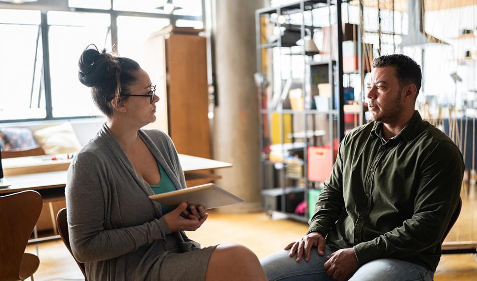 A man and a woman sit on chairs in an office space, facing each other & talking seriously.