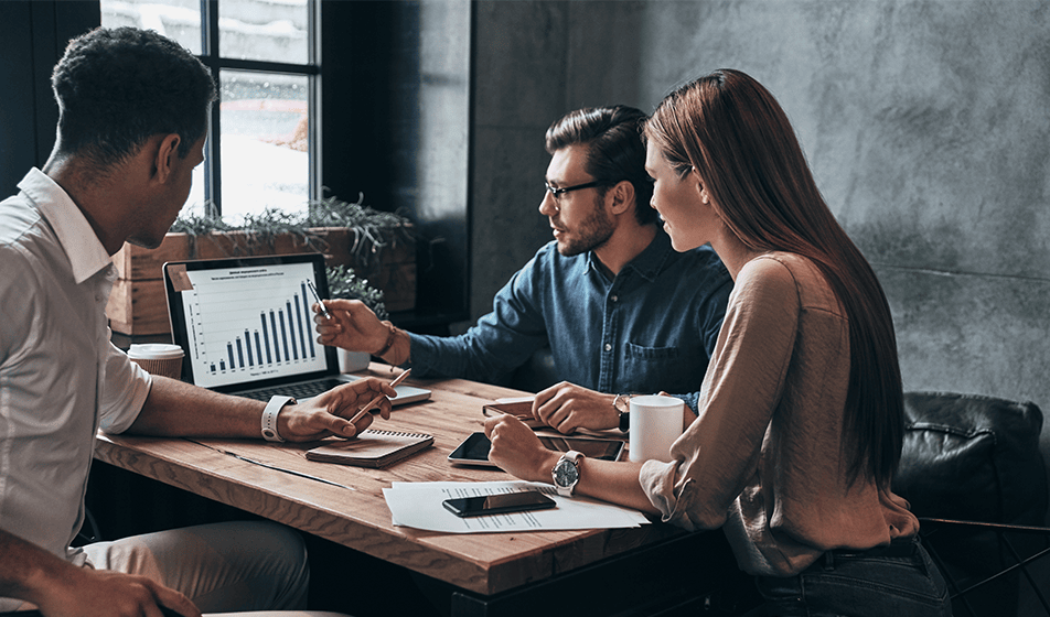 Two men and a woman are in an office, sat around a table, looking away from the camera and at a graph on a laptop. 