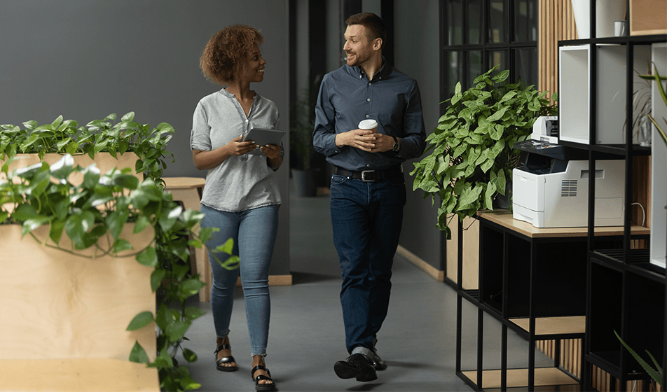 A man and a woman walk through an office environment, talking and looking at each other, holding coffee cups. 