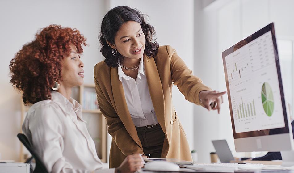 Two women are in an office environment. One is sat at a desk, and the other woman is leaning over and pointing at graphs on the other's screen. Their body language is relaxed and positive. 