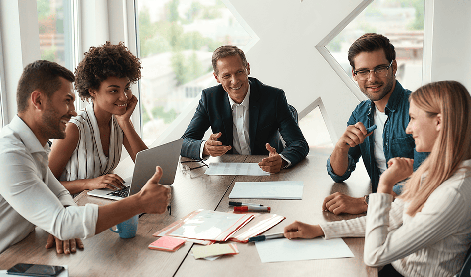 Five colleagues sat around a wooden table and smiling at each other. In front of them is a lot of paperwork, a laptop, stationery and their phones.