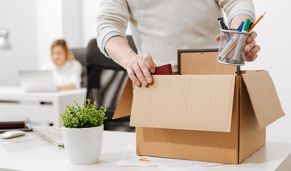 A person in a white jumper has been made redundant. They are filling up a cardboard box with stationery to take home with them.