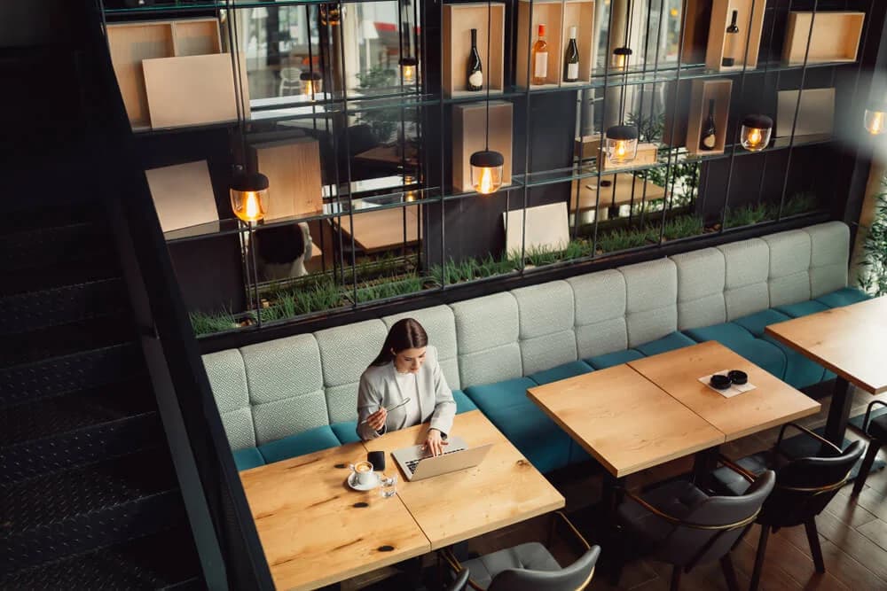 A woman is working remotely from a coffee shop. She is enjoying the the flexible working that her company offers. She is sat at a wooden table with her laptop, phone and a coffee cup in front of her.