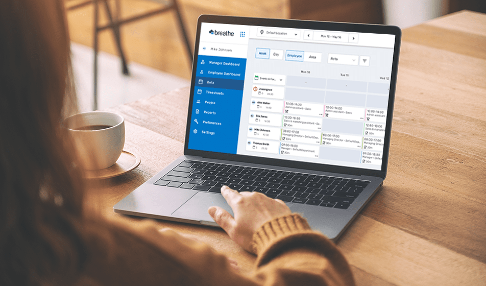 A woman is using a laptop at a wooden desk. On her laptop screen is a display showing Breathe's rota, time and attendance tool.