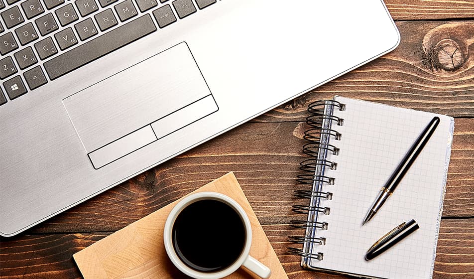 A laptop, cup of coffee and a notepad and pen are shown from above on a wooden desk