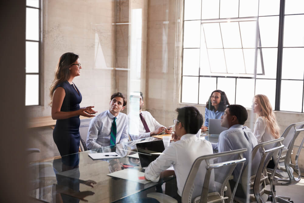 Seven colleagues are having a meeting in a glass-walled office. One colleague is standing up and speaking to the rest. They are all engaged, watching her and listening carefully.