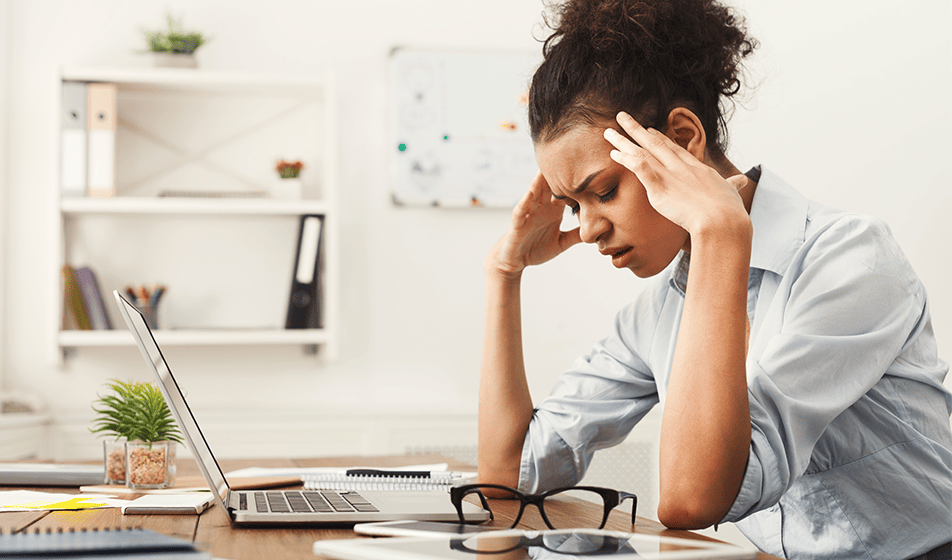 A woman sits at a desk at home in front of her laptop, head in her hands, eyes closed, looking stressed. 