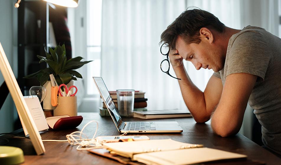 A man is sat at his desk, working from home, resting his head on his hands and looking at his computer screen in slight despair.