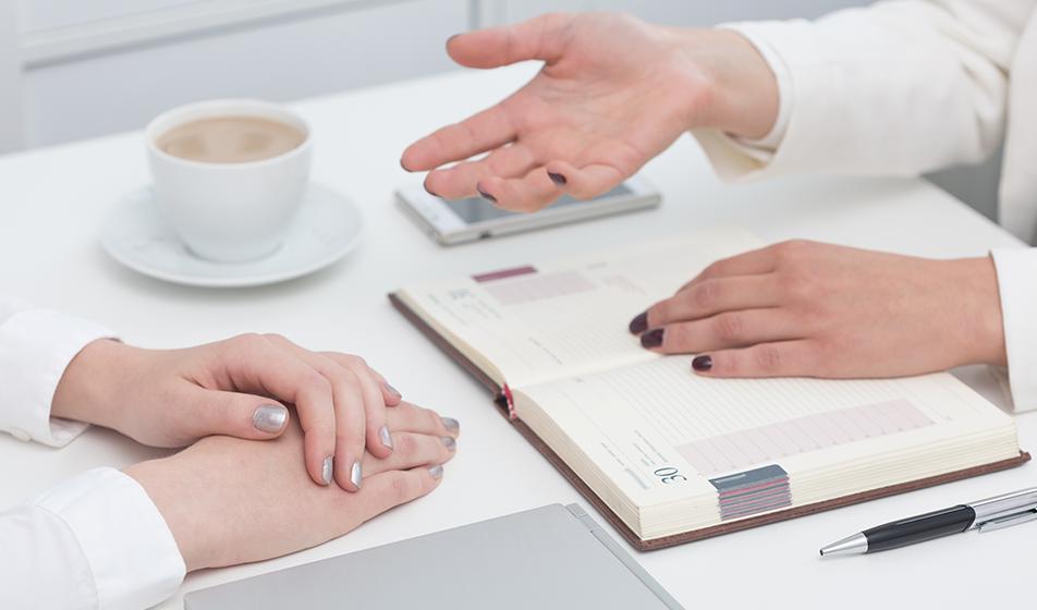 Two pairs of women's hands are shown, sat on either side of a desk. One pair of hands is clasped, the other pair open as if explaining. A tea cup is nearby, and one has a notepad and pen.