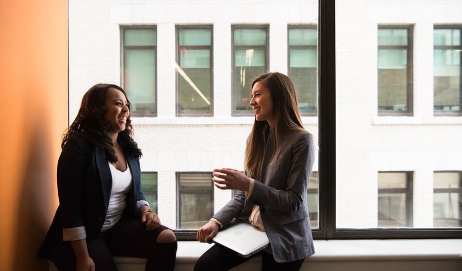 Two female colleagues sat by a window and laughing together. One has her laptop on her lap. 