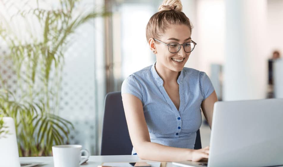 A woman in a blue shirt is working at a desk on her laptop. She has a white mug on her table and has a palm plant behind her.