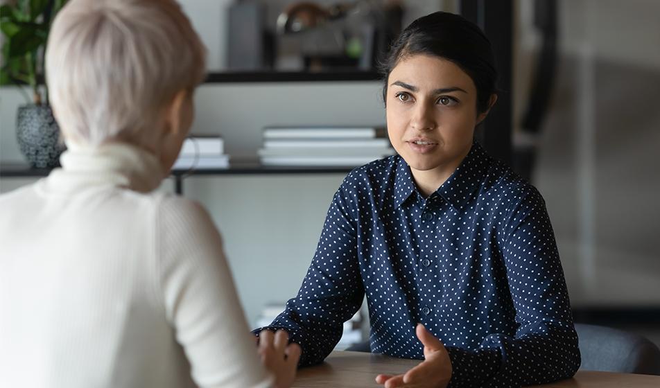 Two women sit across a desk in an office, having a discussion. 