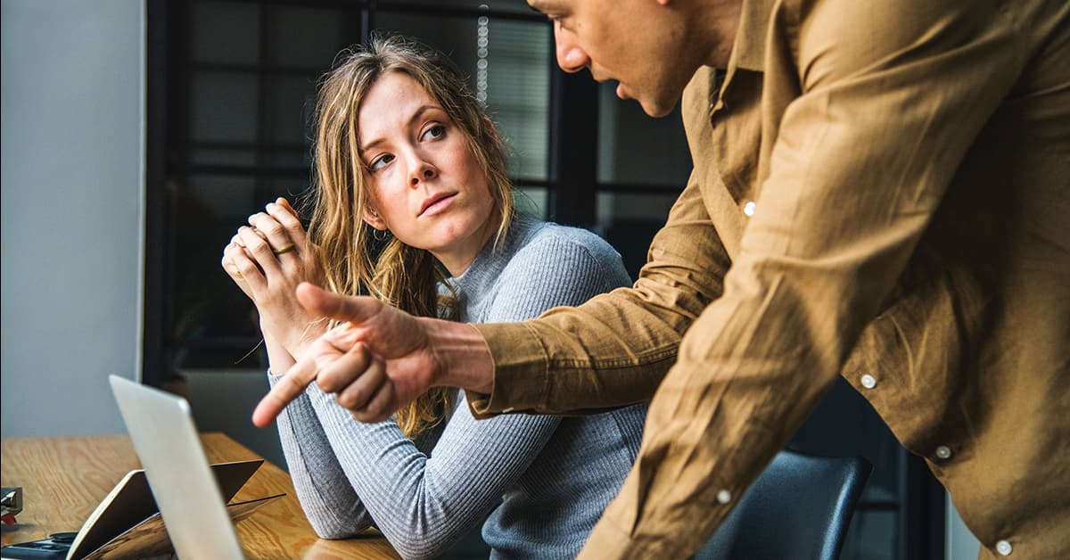 A man in a brown shirt is showing his female colleague something on a laptop. She is watching him explain it to her.