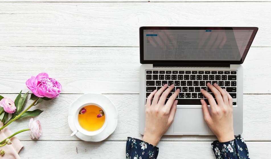 A woman with a blue flowery blouse is typing on her laptop keyboard. She has a cup of tea next to her and some pink flowers on her desk.